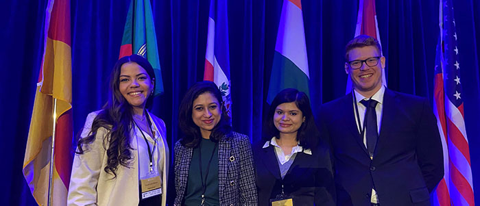 Four business students standing in front of flags and a blue curtain