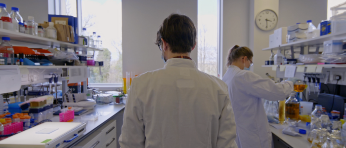 Man wearing lab coat walking between two lab benches.
