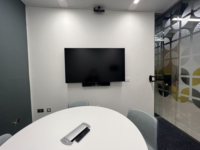 Flat-floored meeting room with round table and chairs, and wall mounted video monitor.