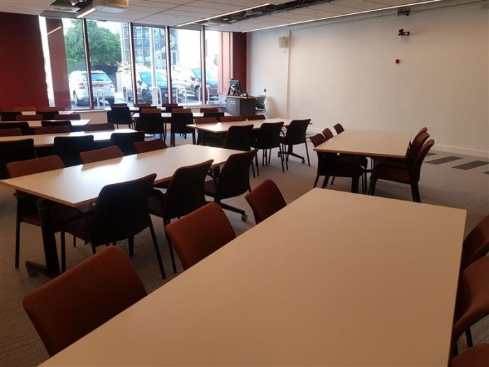 Flat floored teaching room with tables and chairs, projector, videoconferencing mics and camera, PC, and lectern. 
