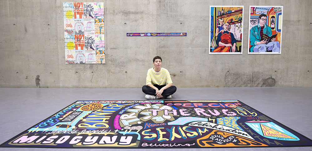 Artist Molly Hankinson sits on floor behind a large colourful mural. Two artworks hang on wall behind her.