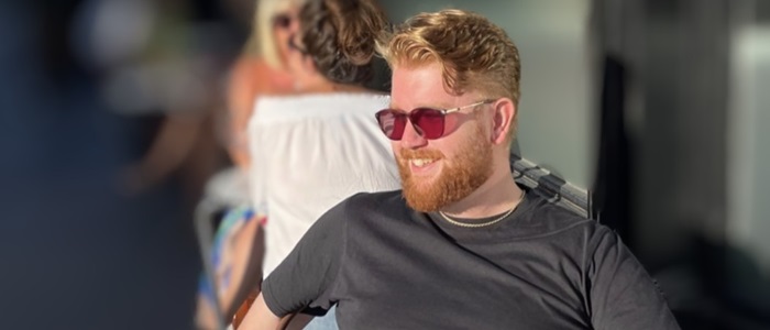 Man with red hair, a beard and sunglasses wearing a grey t-shirt looking and smiling to the side