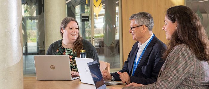 Three staff members sitting talking at a table in front of open laptops