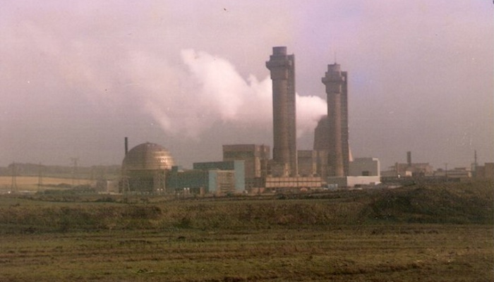 A view of the buildings of Sellafield nuclear power station with steam plumes emitting from it, seen across a field and against a stormy sky