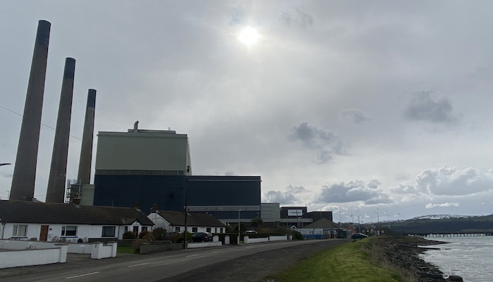 A power station with three tall thin chimneys and a square building, with small white cottages and a bay of the sea in front of it. Photo credit: Ewan Gibbs
