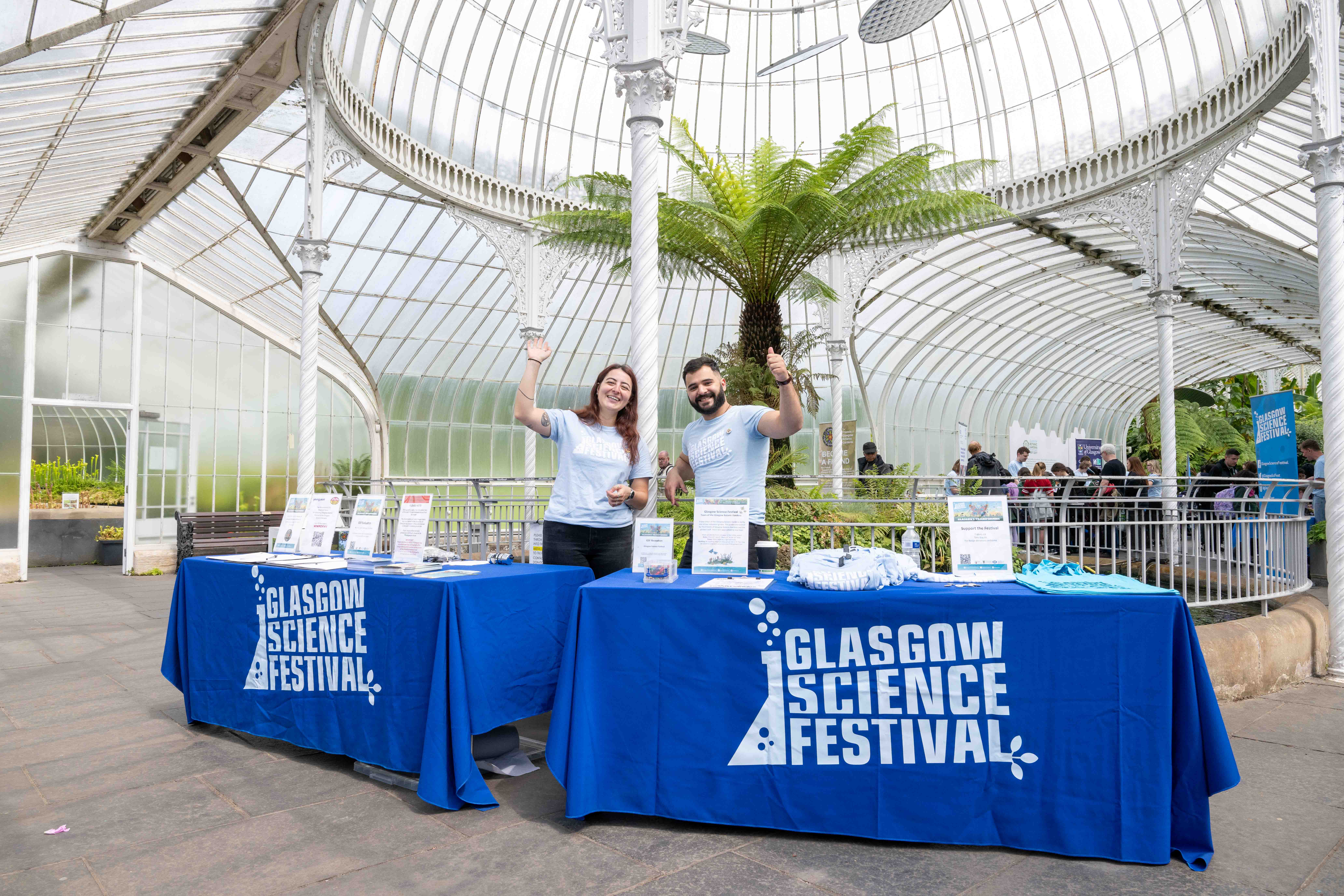 Two people in the Glasgow Botanic Gardens wearing blue T-shirts. Standing behind two tables with tablecloths that have the Glasgow Science Festival logo. The people are waving to the camera.