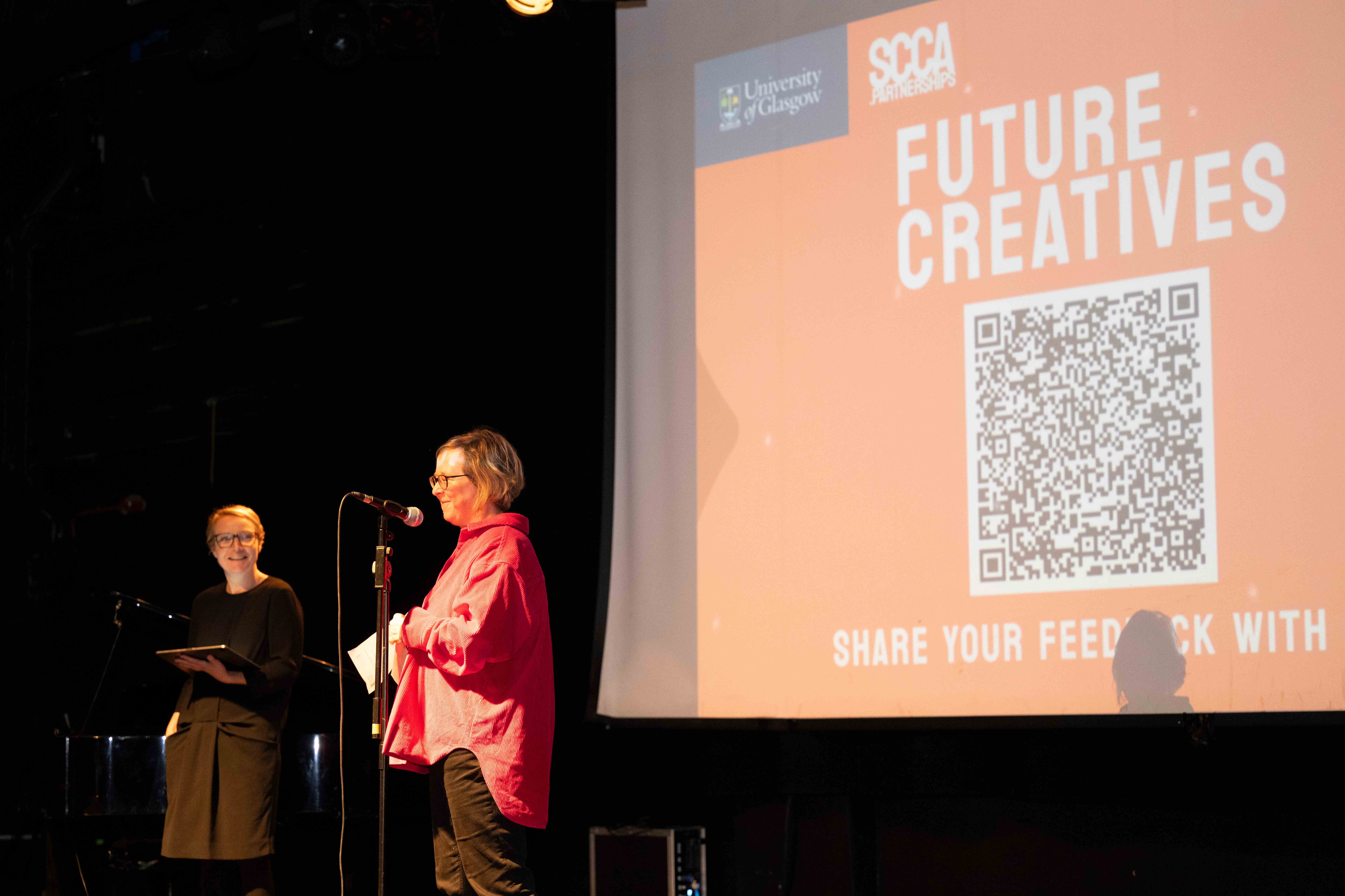Two white women stand in front of a screen projecting text reading