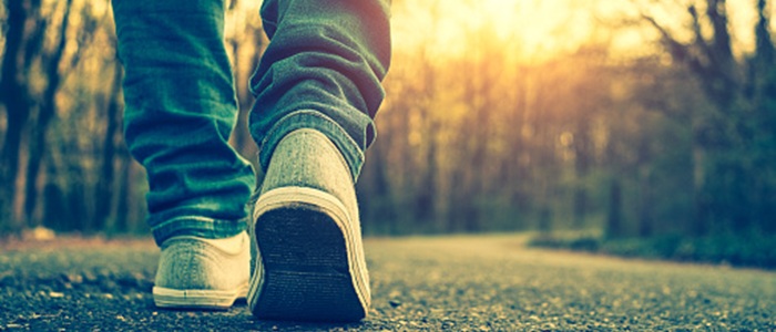 Person wearing jeans and trainers walking along a road. There are trees both sides and low sunlight.