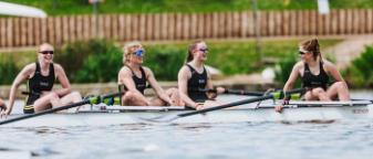 Team of four woman's rowing team in boat on water. Looks like they have just finished a race and are happy.