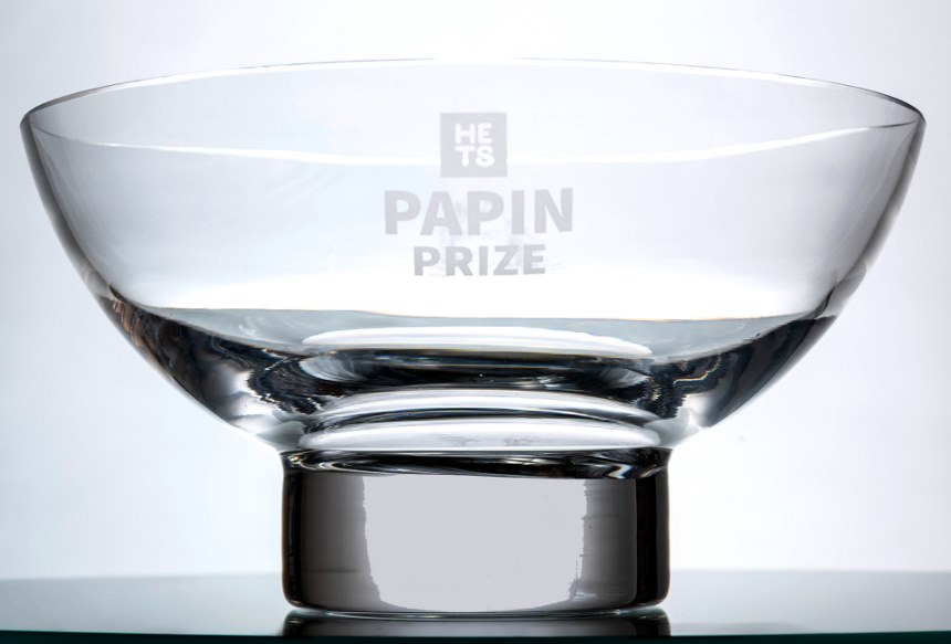 A close up image of a trophy bowl, which is clear, with a silver base, and the words HETC Papin Prize etched on the glass