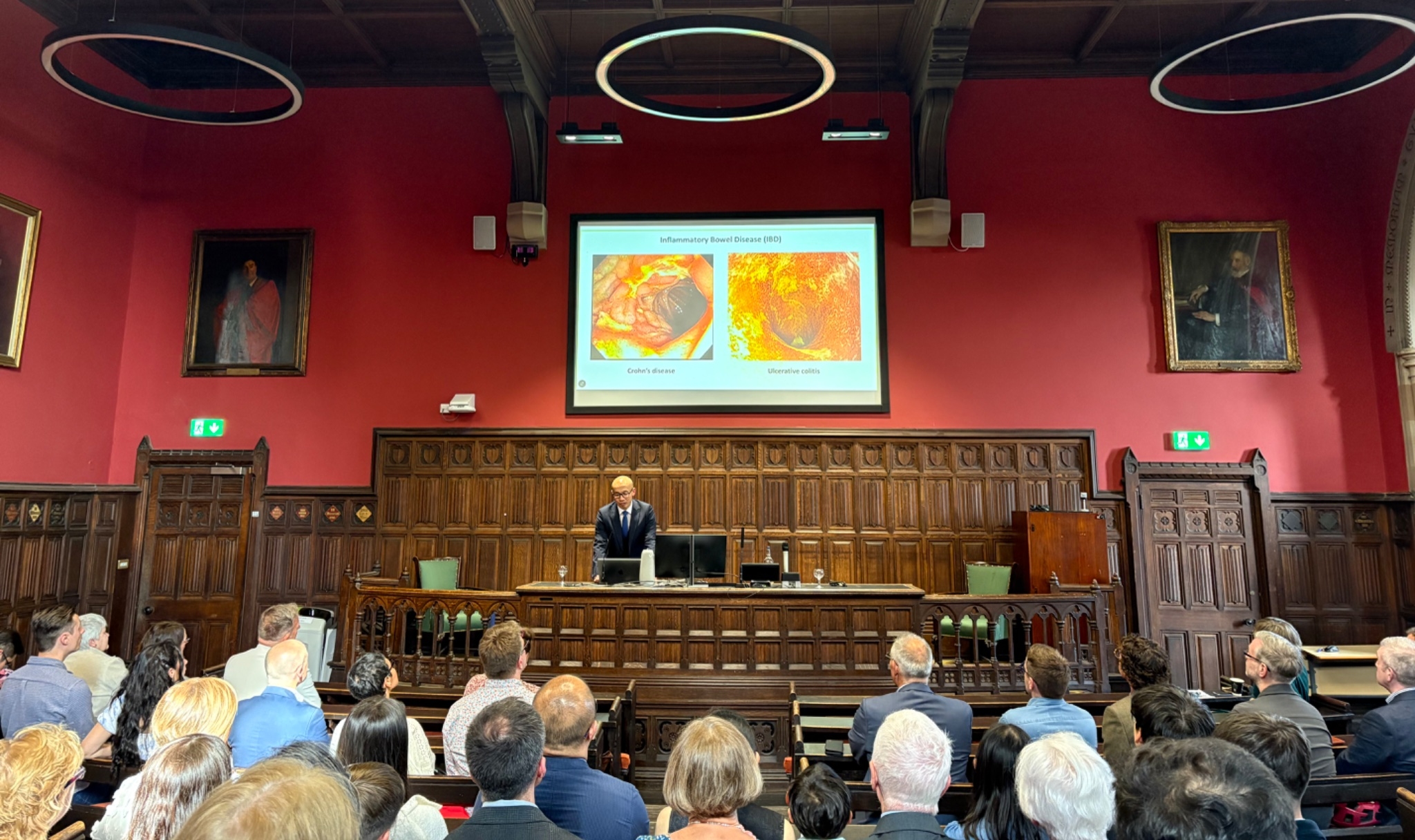 A wide-angle shot showing Professor Ho at the podium and the presentation screen, taken from further back in the room showing the engaged audience