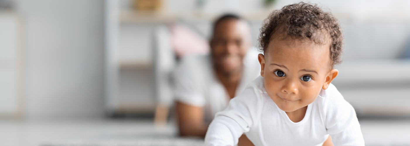 A baby is crawling towards the camera with dad lying on the floor behind