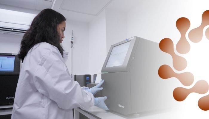 Researcher in a white lab coat operates a DNA sequencing machine in a genomics laboratory with modern equipment and screens.
