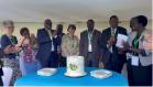 A group of formally dressed people stand around a table with a decorated white cake featuring green and brown accents. Stacks of plates are placed beside the cake, and the individuals are wearing lanyards, indicating they are attending an event or celebration.