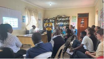 A group of people attending a community meeting in a classroom-like setting, with one person presenting information projected on a screen.