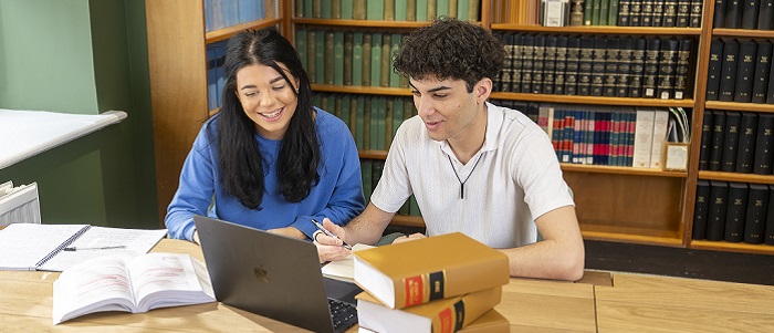 An image of two students working together at a desk with a laptop on the table, and bookshelves behind them.