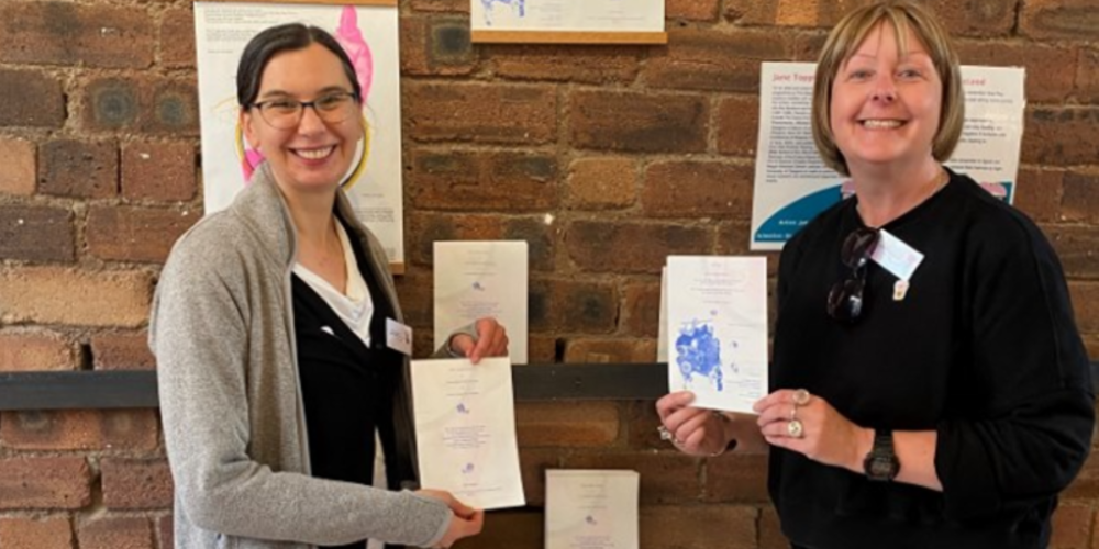 Dr Megan MacLeod and Jane Topping stood in front of an indoor brick wall, holding up paper to show the science-inspired art