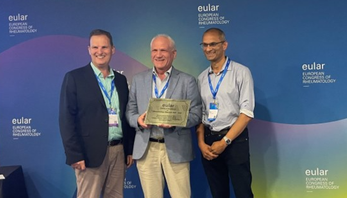 Image (from left to right): Professor Iain McInnes (Head of College of MVLS), Ronald Van Vollenhoven (Research Chair of EULAR), Professor Neil Basu (Sii Professor of Musculoskeletal Medicine & Vasculitis) stood together holding a plaque in front of a screen with EULAR branding