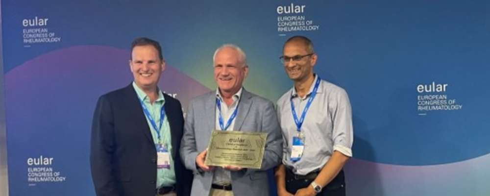 Image (from left to right): Professor Iain McInnes (Head of College of MVLS), Ronald Van Vollenhoven (Research Chair of EULAR), Professor Neil Basu (Sii Professor of Musculoskeletal Medicine & Vasculitis) stood together holding a plaque in front of a screen with EULAR branding