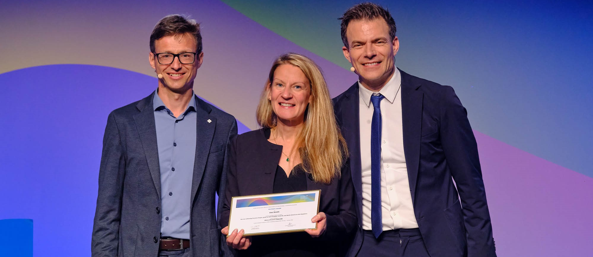 Dr Eve Smith stood on the stage at the EULAR Conference holding a certificate of her award, with those presenting the award stood on either side
