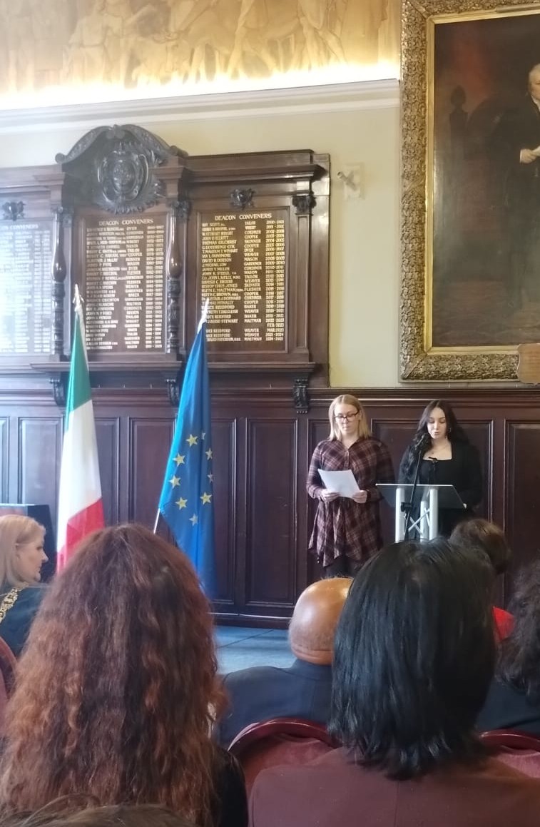 Carys Bilton and Eva Merritt stand behind a podium and give a reading to an assembled crowd. An Italian and an EU flag stand next to them.