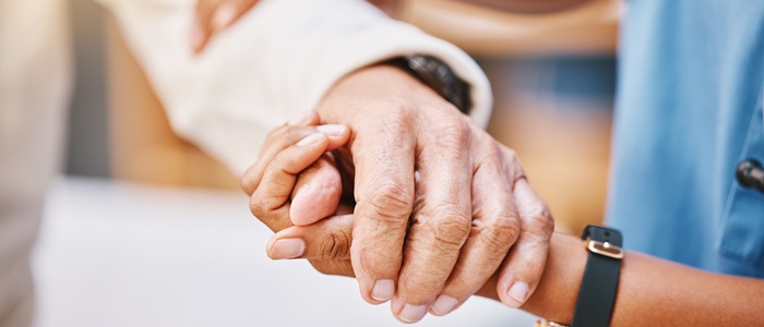 A patient is holding the hand of a health professional who is wearing a blue tunic