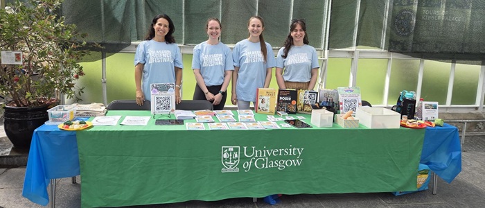 Four researchers wearing blue Glasgow Science Festival t-shirts standing behind a table. The green tablecloth has the University of Glasgow logo on it.