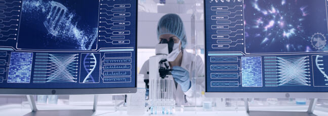 A scientist dressed in PPE is looking at something under a microscope. There are computer screens with images of a dna helix