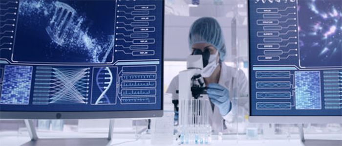 A scientist dressed in PPE is looking at something under a microscope. There are computer screens with images of a dna helix