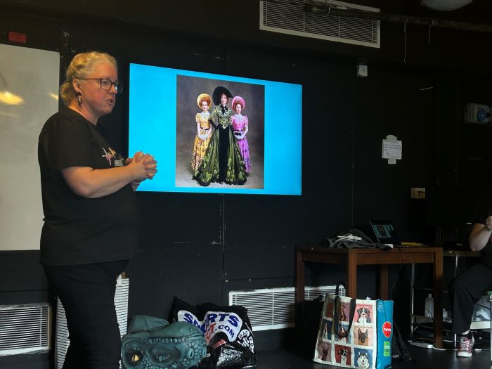 A woman standing speaking in front of a screen displaying historic womens clothing