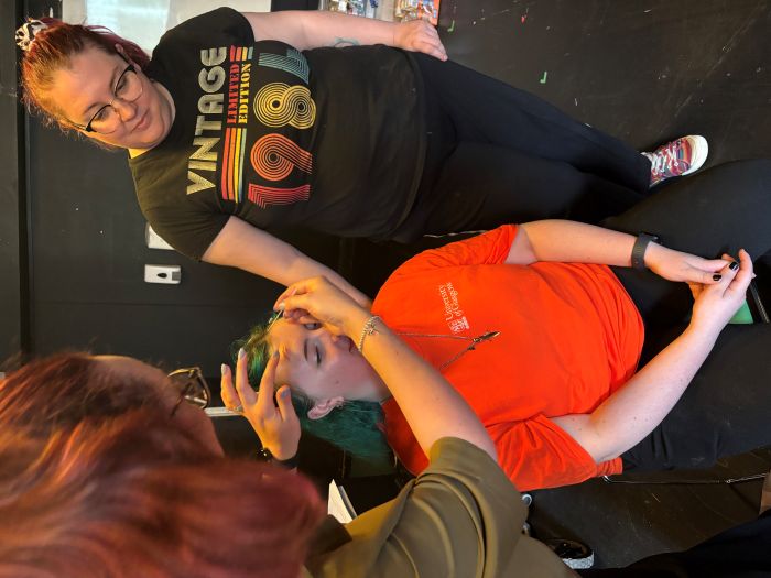 one person sits on a chair while two other people put makeup on them