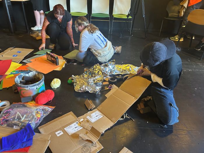 Young people sit on the floor making props out of various materials including carboard, yarn and paper