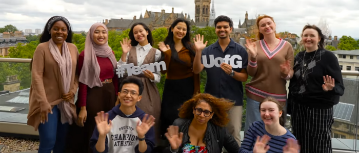 A group of students standing on the JMS balcony with the Gilbert Scott tower in the background, the students are waving at the camera and holding Team UofG signs