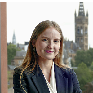 Profile photo of  Tuuli Vanhapelto standing at a window with the view of the University of Glasgow tower in the background