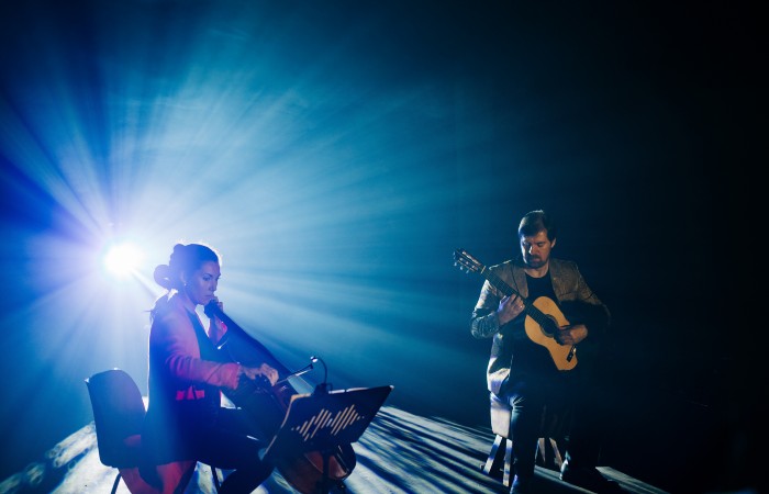 A musician playing cello and a musician playing guitar sit next to each other on a stage in front of a bright stage light