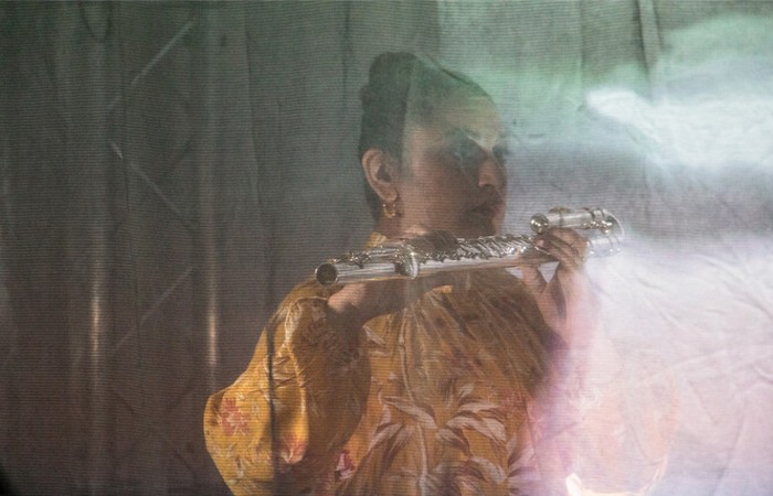 Image shows Diljeet standing, holding a flute to her face. She is wearing a yellow floral patterned outfit.