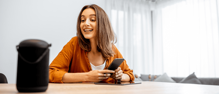 Woman holding mobile phone and addressing a smart speaker on the table in front of her
