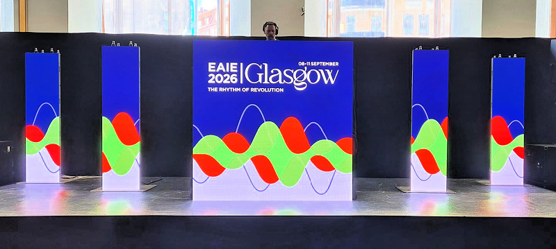 DJ bstood behind DJ booth with panels on the booth that say 'Glasgow 2026, The rhythm of revolution'