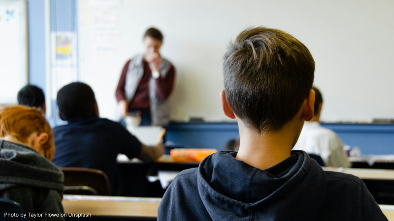 Photo of boy sitting in classroom with teacher in background