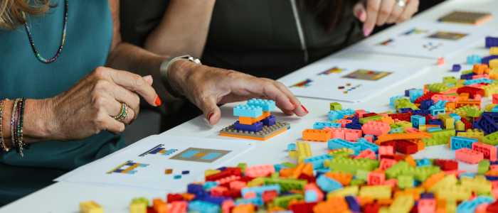 a large amount of colourful lego is on a white table. A person is sitting at the table - only their hands are visible as they build a blocky structure on a baseplate