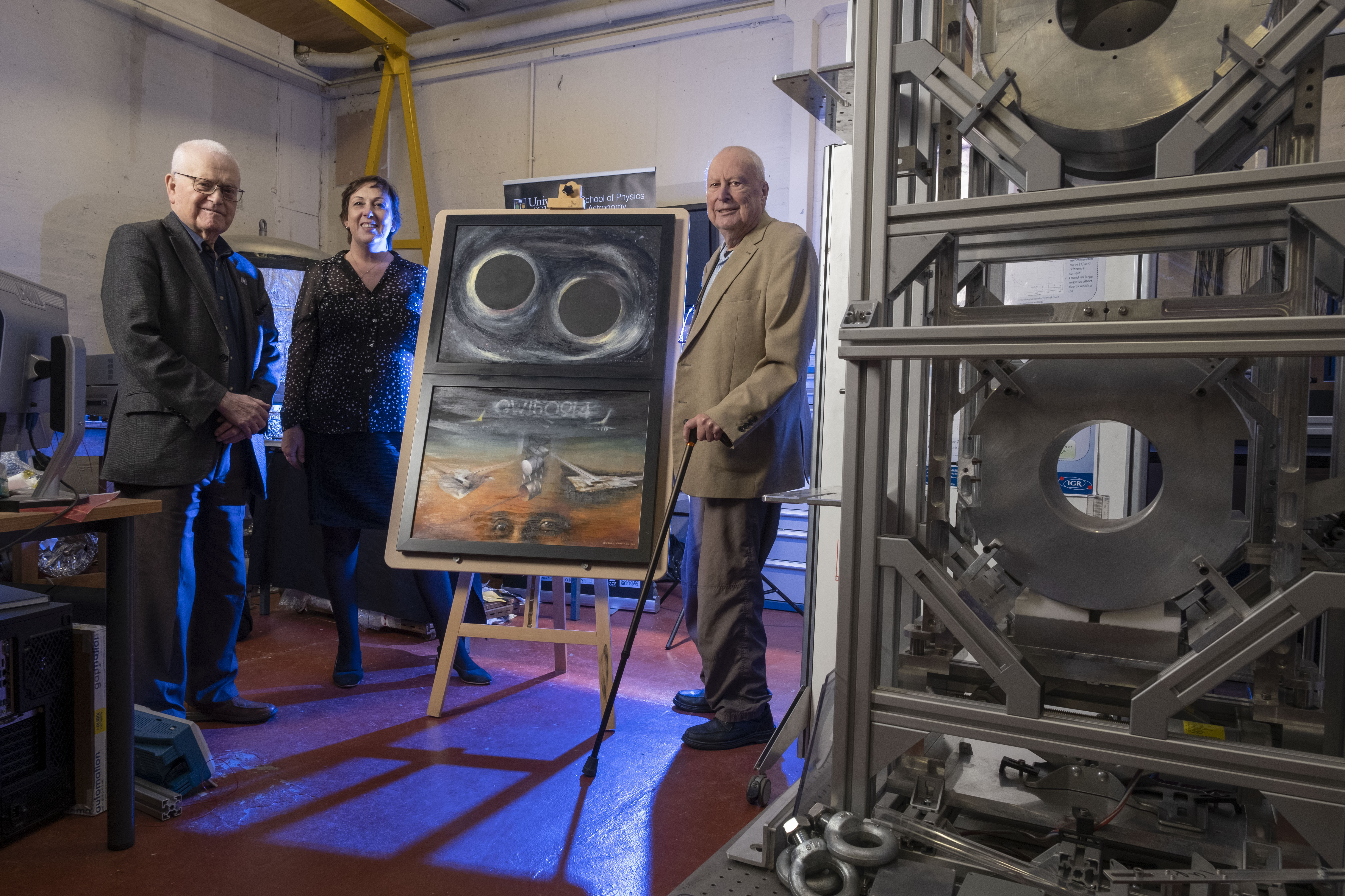 Professor Dugald Cameron; Professor Sheila Rowan, director of the University of Glasgow’s Institute for Gravitational Research, and Professor Sir James Hough pose with Professor Cameron’s artwork alongside a model of the mirror suspensions developed and built at the University of Glasgow which made LIGO’s historic first detection of gravitational waves possible