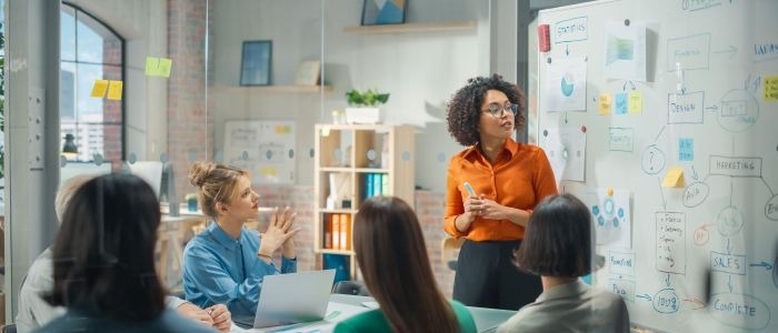 A group of women gather in a meeting room. One women is speaking next to a white board covered in text.