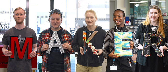 Five people standing in a line holding each letter of the word Maker - each letter is made up of different materials