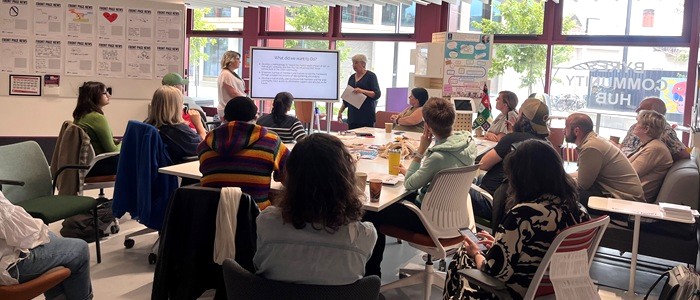 A group of people sitting round a big table in Byres Community Hub. They are listening to a speaker who is presenting information on a screen