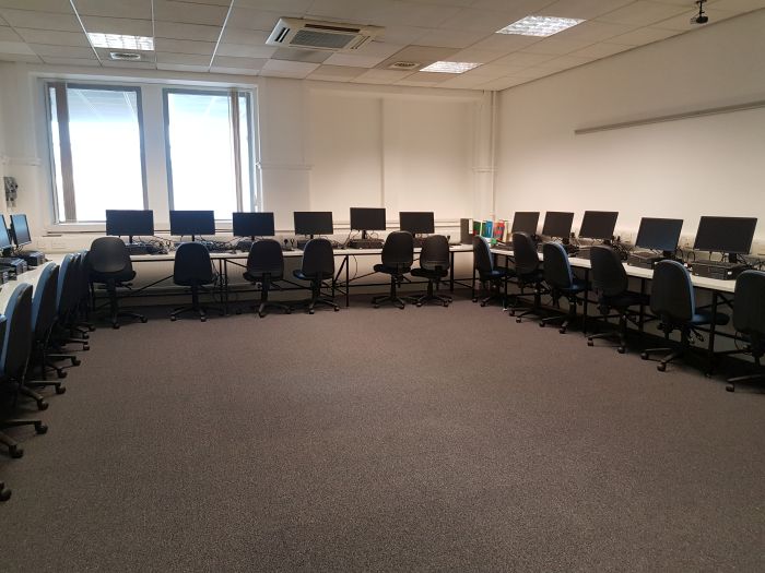Flat floored IT lab with computer stations positioned around perimeter of the room, each consisting of a PC with monitor, and a chair.