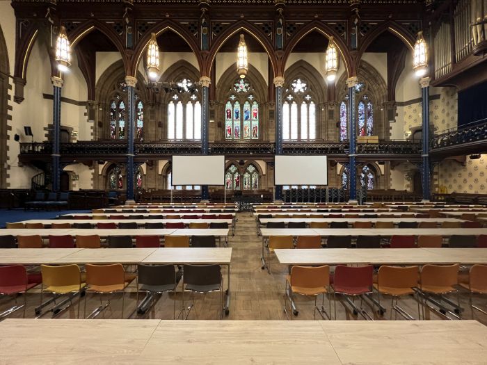 Flat floored hall with tables and chairs in rows, and large screens.