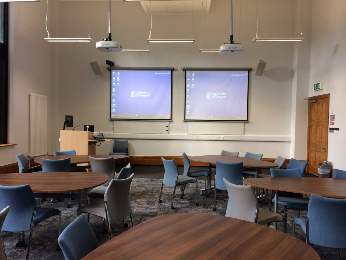 Flat floored teaching room with groups of teardrop tables and chairs, projectors, screens, and PC