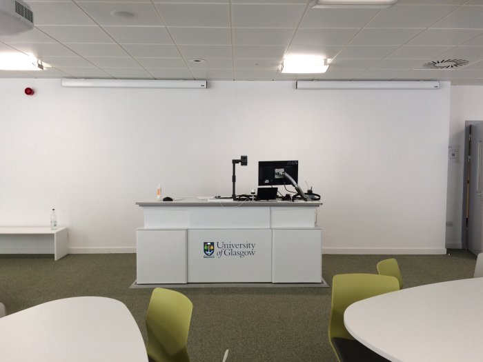 Flat floored teaching room with groups of teardrop tables and chairs, projectors, PC, and lectern.