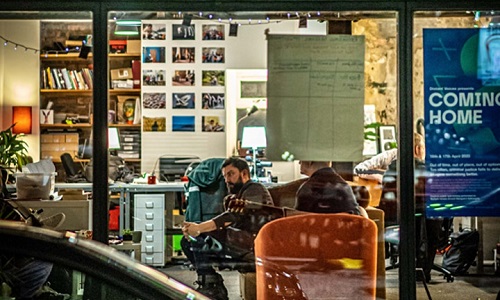Various people sitting inside a brightly lit studio, viewed from outside through the glass