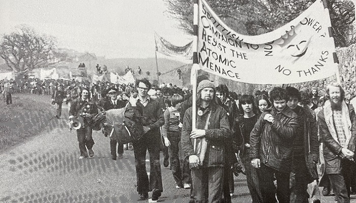 A black and white photo of a demonstration with people carrying a banner that says 'Scottish Campaign to Resist the Atomic Menace' and 'Nuclear Power? No Thanks' with a sun with a smiley face in middle. Source: 'Torness: Nuclear Power Station: From Folly to Fiasco (Edinburgh: SCRAM, 1983)'